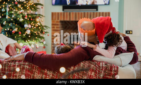 Father is curled up on the sofa ith his daughter at christmas time, watching television. Stock Photo