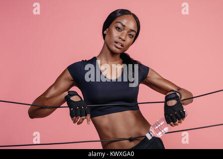 Muscular young woman with rubber bands in studio. Female standing with elastic band and water bottle on pink background. Stock Photo