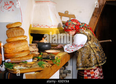Woman in Ukrainian native costume puts pot with traditional device rogach in a rural oven, vintage rural house. March 23, 2018. Kyiv, Ukraine Stock Photo