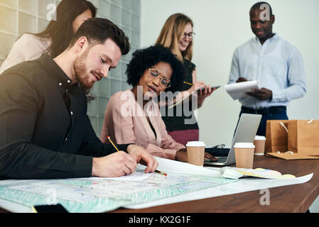 Two designers discussing building blueprints together while working at a table in an office with colleagues in the background Stock Photo