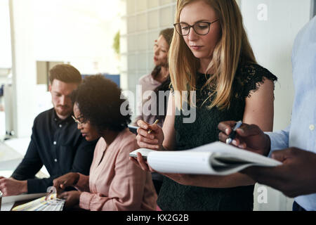 Two coworkers writing down notes on clipboards while working together in a modern office with colleagues in the background Stock Photo