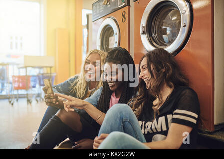 Three young female friends laughing and sitting together on a laundromat floor using a cellphone Stock Photo