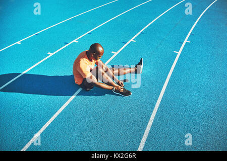 Young African male athlete in sportswear sitting alone on the lanes of a running track tying up his running shoes before training Stock Photo