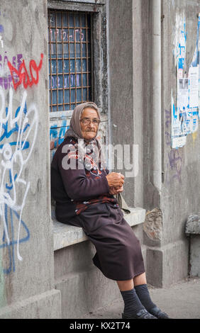 Local lifestyle: poor old woman sits on a window ledge with graffiti begging from passers-by in Bucharest, capital city of Romania, central Europe Stock Photo