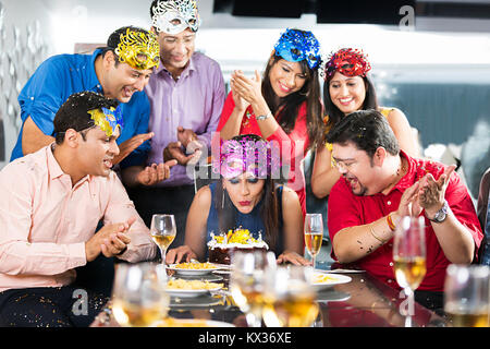 Adult woman blowing out candles on her cake at-birthday party surrounded by celebrating Stock Photo