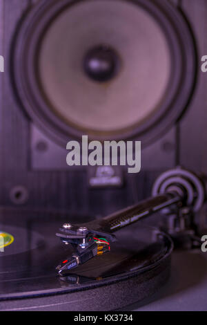 Vinyl record on a turntable, with tonearm and stylus on the LP, and a single black speaker cabinet from a stereo hi-fi sound system, standing behind. Stock Photo