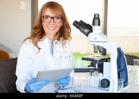 Shot of a middle aged scientist woman using digital tablet while sitting at desk in front of microscope and working on new medical research. Stock Photo