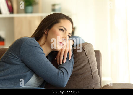 Sad woman looking trough a window sitting on a couch in the living room at home Stock Photo