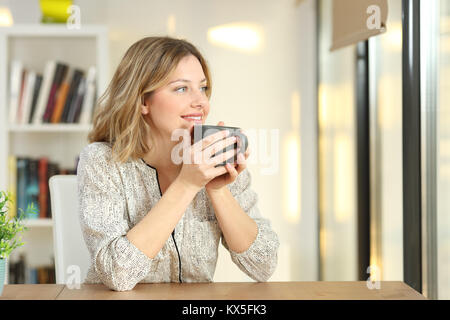 Portrait of a woman looking through a window drinking coffee sitting in a table at home Stock Photo