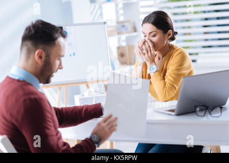 Ill woman closing her eyes while sitting at the table and sneezing Stock Photo