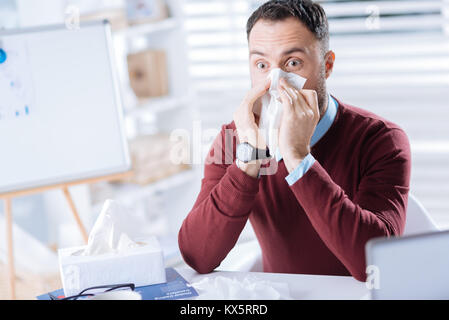 Emotional man blowing his nose while being at work alone Stock Photo