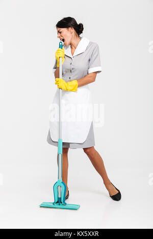 Full length portrait of young maid in gray uniform having fun while singing in mop as microphone, isolated on white background Stock Photo