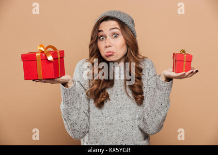 Funny confused brunette girl in woolen hat and jersey holding two gift boxes, looking at camera, isolated on beige background Stock Photo