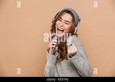 Close-up portrait of funny brunette girl in headphones holding her smartphone like microphone and singing, isolated on beige background Stock Photo