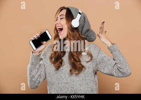 Portrait of emotional young woman in gray woolen hat holding her smartphone like microphone and singing, isolated on beige background Stock Photo