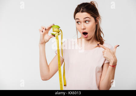 Portrait of an excited pretty girl holding half eaten apple with a measuring tape and pointing finger isolated over white background Stock Photo