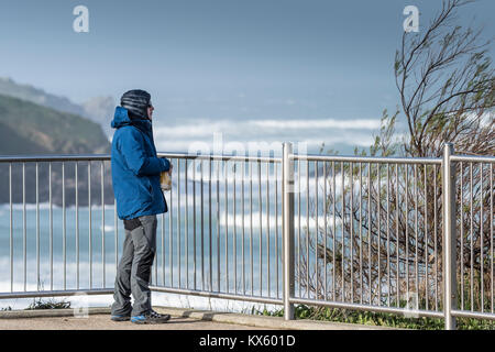 A man eating a snack and looking out to sea wearing warm clothes against the cold windy weather Stock Photo