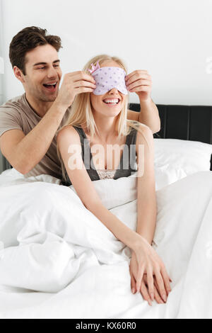 Funny handsome man wearing his blonde cheerful wife sleeping mask while they lying in bed Stock Photo