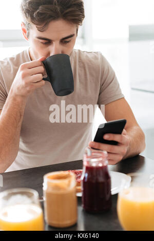 Portrait of young focused man drinking tea and using smartphone while have breakfast Stock Photo