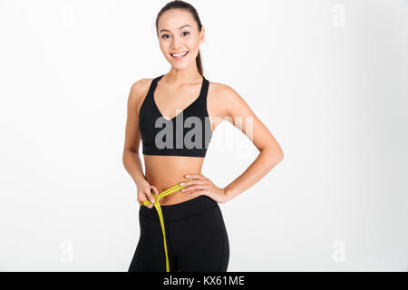 Portrait of a satisfied asian fitness woman holding measuring tape around her waist isolated over white background Stock Photo