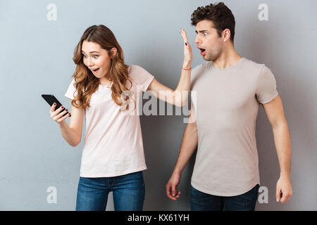 Portrait of a young couple standing with mobile phone, girl holding mobile phone while frustrated man standing near isolated over gray background Stock Photo