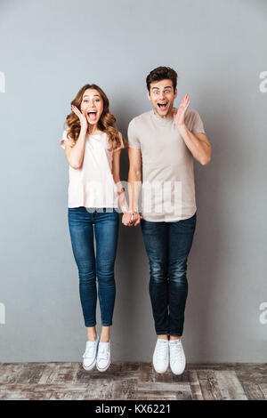 Full length portrait of an excited young couple holding hands and jumping over gray wall Stock Photo