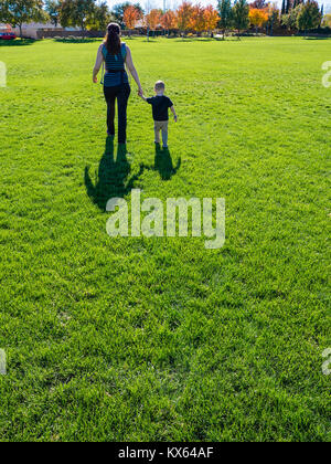 Mother and two-year old son walking together holding hands, walking away, casting long shadows, on green grass in a park. Stock Photo
