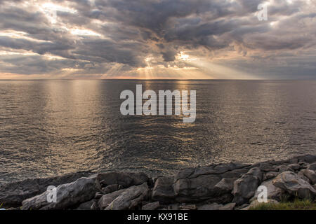 Sun rays shining through dark storm clouds over the ocean with rocks in the foreground. Taken on Cape Breton Island, Nova Scotia, Canada Stock Photo