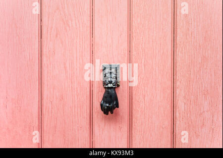 Front view of a black hand-shaped door-knocker fitted on an old pink front door. Stock Photo