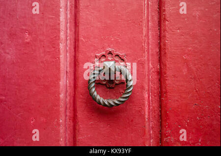 Front view of a round and twisted cast iron door knocker fitted on an old wooden door painted in red. Stock Photo