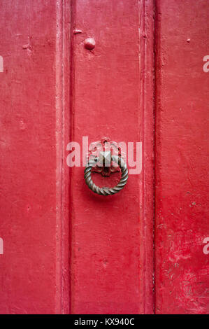 Front view of a round and twisted cast iron door knocker fitted on an old wooden door painted in red. Stock Photo