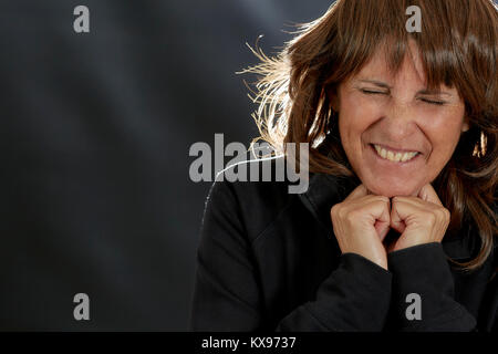 Attractive Middle aged woman with layered brown hair holding her hands under chin with eyes closed and making a distasteful expression with back light Stock Photo
