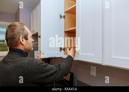 The worker sets a new handle on the white cabinet with a screwdriver installing kitchen cabinets Stock Photo