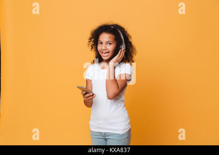 Portrait of a smiling little african girl listening to music with headphones while holding mobile phone and looking at camera isolated over orange bac Stock Photo