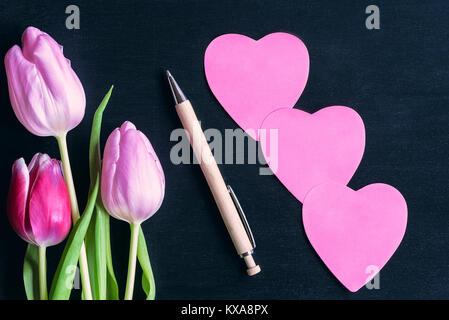 Colorful tulips, a wooden pen, and three pink heart shaped paper post-its, with space for messages, on a black wooden background. Stock Photo