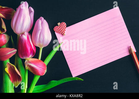 Beautiful colorful tulips, a blank red notebook sheet with stripes and a wooden clip with a heart shape on it, placed on a black wooden background. Stock Photo