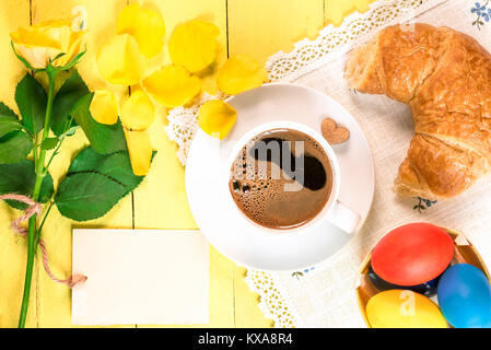 Breakfast table with a cup of coffee, freshly baked croissant, painted Easter eggs and a yellow single rose with fallen petals and a message card. Stock Photo