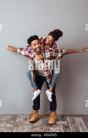 Full length portrait of a smiling young african man holding pretty girfriend on his back and having fun isolated over gray background Stock Photo
