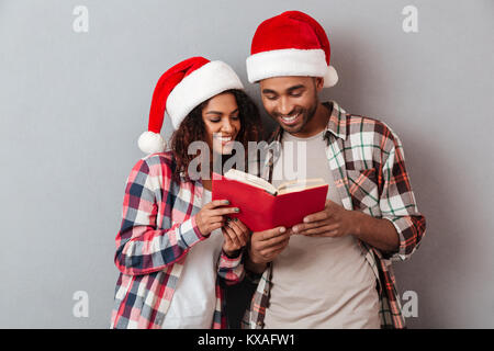 Portrait of a happy young african couple dressed in christmas hats reading a book while standing isolated over gray background Stock Photo