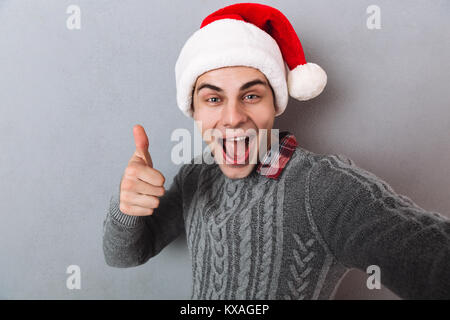 Cheerful Man in sweater and christmas hat making selfie and showing thumb up over gray background Stock Photo