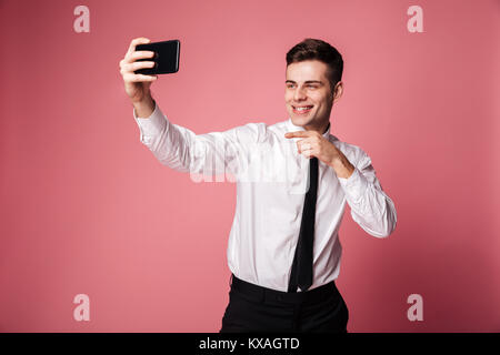 Photo of funny young businessman standing isolated make selfie by mobile phone. Looking aside pointing. Stock Photo