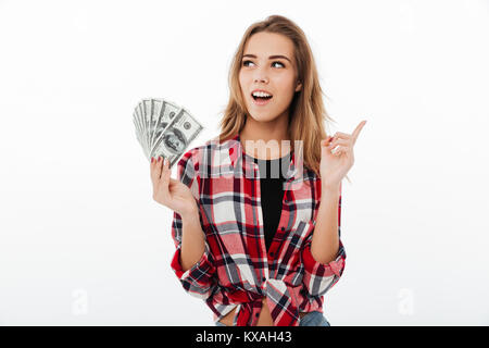 Portrait of an excited smiling girl in plaid shirt holding bunch of money banknotes and pointing finger up isolated over white background Stock Photo