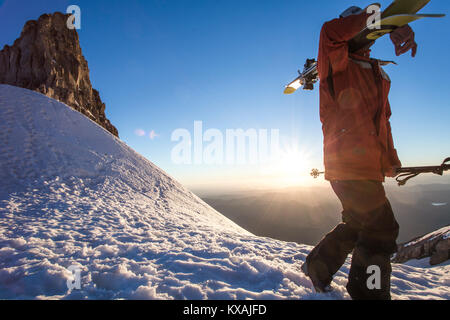 Skier carrying skis on snowy mountain with Sun setting in background, Mount Hood, Oregon, USA Stock Photo