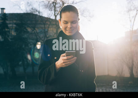 Young smiling woman in a black coat looks in her mobile phone outdoors Stock Photo