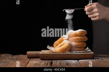 Baker sprinkles sweet donuts with powder sugar on black background. Delicious, but unhealthy food on the old wooden table with copy space. Stock Photo