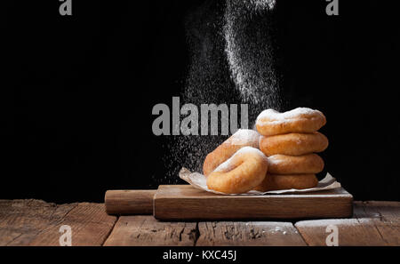 Baker sprinkles sweet donuts with powder sugar on black background. Delicious, but unhealthy food on the old wooden table with copy space. Stock Photo