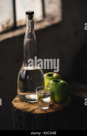 Apple brandy, rustic retro atmosphere. Bottle and single shot glass with alcohol drink and two green apple fruit. Stock Photo