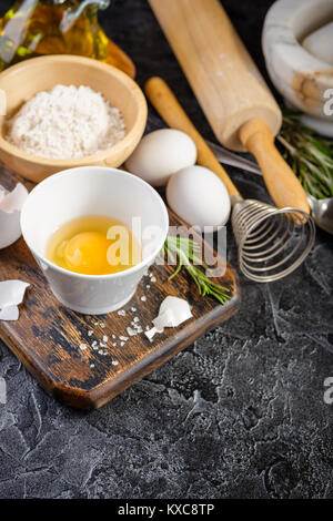 Raw uncooked flour, eggs, rosemary, salt and olive oil on wooden cutting board over dark background. Stock Photo