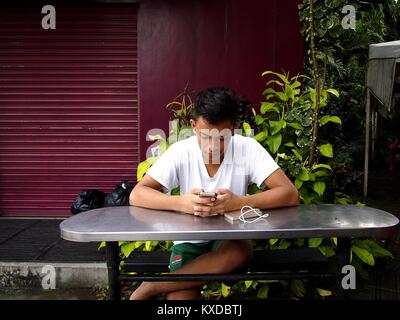 ANTIPOLO CITY, PHILIPPINES - DECEMBER 28, 2017: A young man uses his smartphone while sitting at a table at public park. Stock Photo