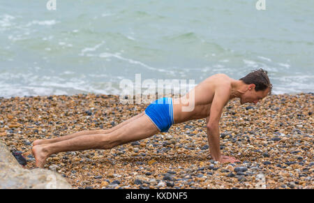 Man wearing only shorts doing morning exercises (pushups, push ups, pressups, press ups) on a beach on a cold day in the UK. Stock Photo